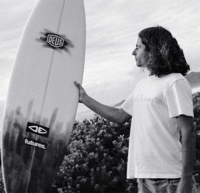 Eddy holding his surfboard that he hand shaped. He is standing infront of a beach break